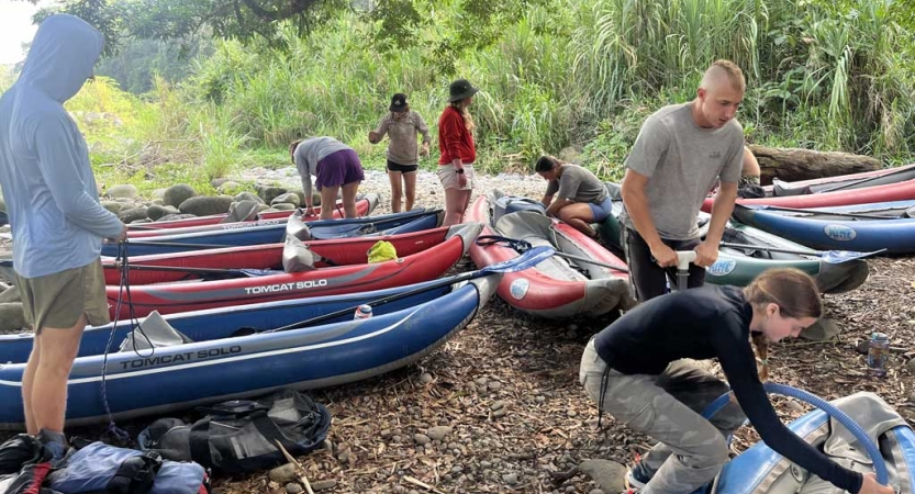 several watercraft rest on the ground while people appear to be getting things read in and around them.